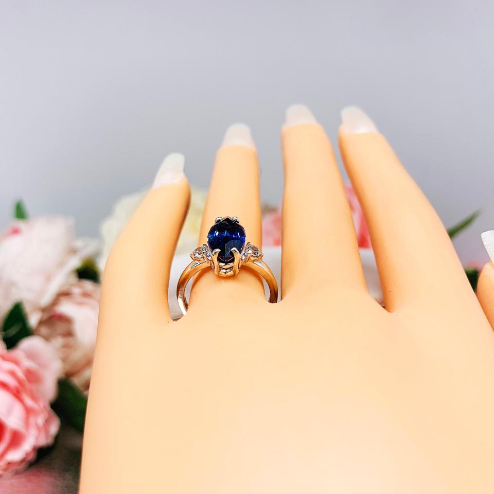 Mannequin hand with long white shimmery nails, displayed against a soft white-gray background with pink and white floral accents. The hand is wearing a sterling silver ring showcasing a striking marquise-cut lab-created blue sapphire, it is flanked by two brilliant round cubic zirconia stones.