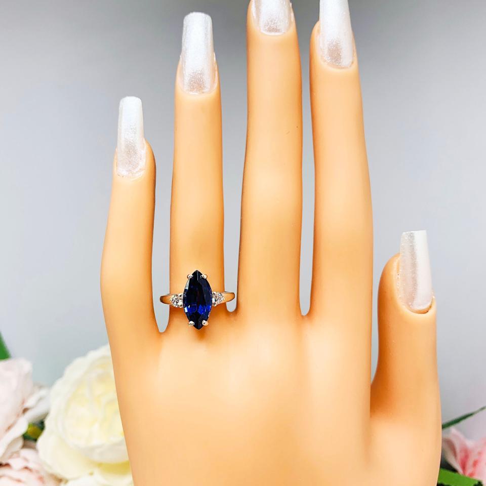 Mannequin hand with long white shimmery nails, displayed against a soft white-gray background with pink and white floral accents. The hand is wearing a sterling silver ring showcasing a striking marquise-cut lab-created blue sapphire, it is flanked by two brilliant round cubic zirconia stones.