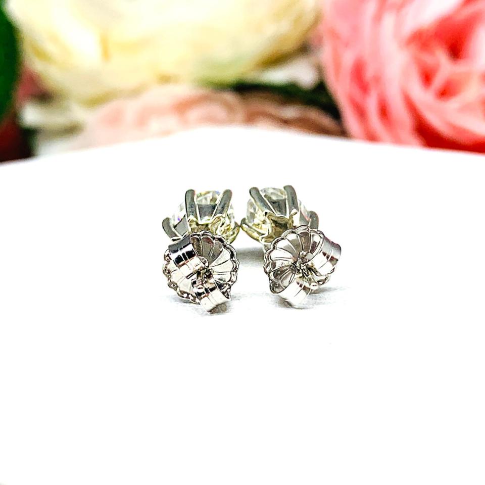 A pair of 10k whtie gold stud earrings set with oval brilliant cut moissanites.  The 6 prong earrings are on a white surface with both earrings facing toward the back to showcase the center post and butterfly friction clasps.