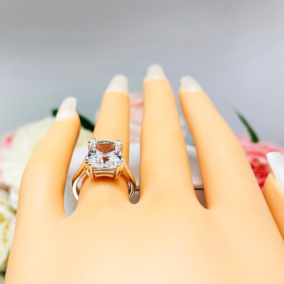 Mannequin hand with long white shimmery nails, displayed against a soft white-gray background with pink and white floral accents. The hand is wearing an elegant sterling silver cathedral engagement ring set with a large radiant cut cubic zirconia.