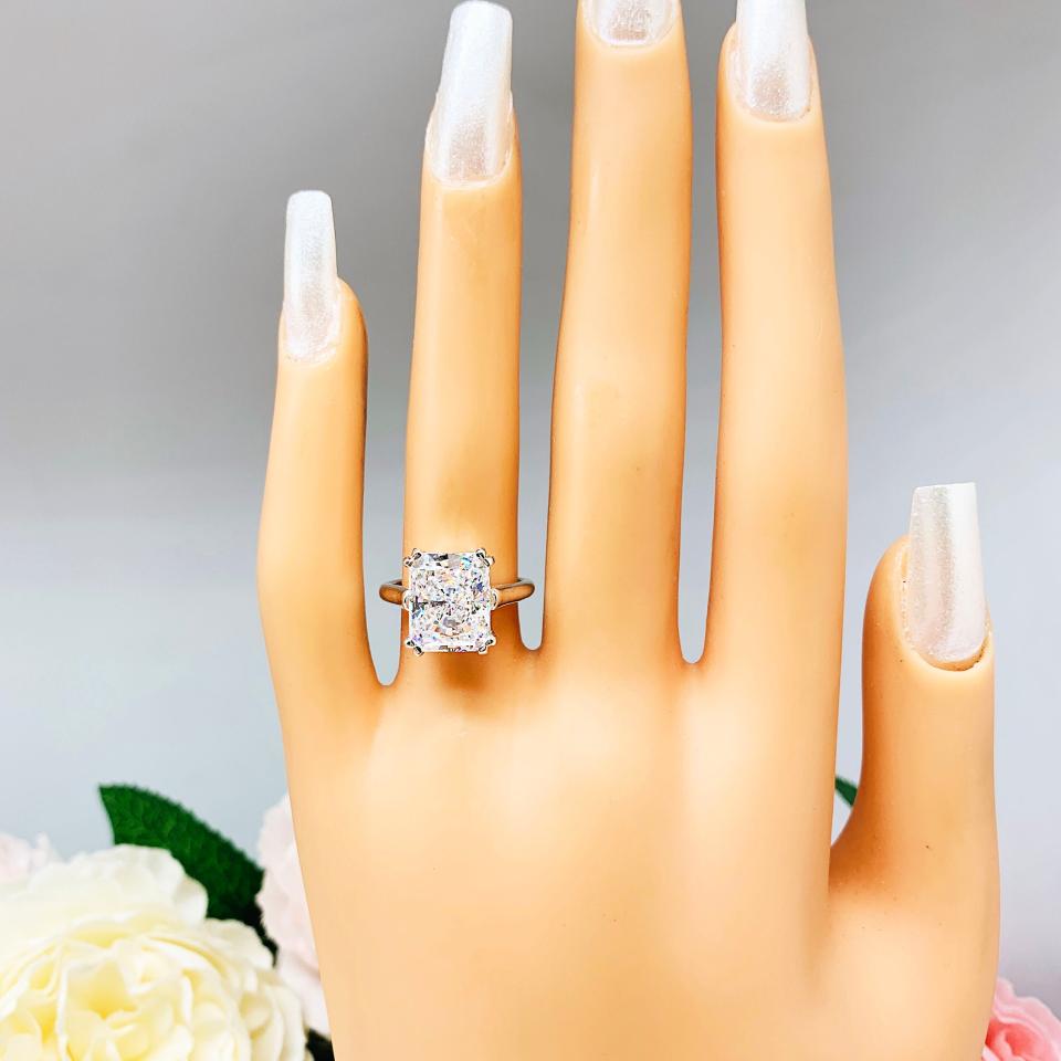 Mannequin hand with long white shimmery nails, displayed against a soft white-gray background with pink and white floral accents. The hand is wearing an elegant sterling silver cathedral engagement ring set with a large radiant cut cubic zirconia.