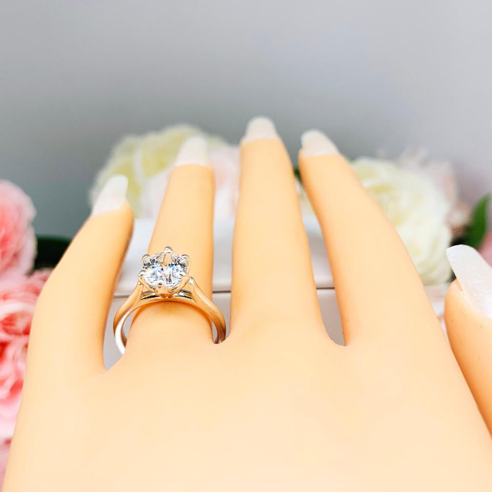 Mannequin hand with long white shimmery nails, displayed against a soft white-gray background with pink and white floral accents. The hand is wearing a sterling silver engagement ring with an oval lab created white sapphire set in a 6-prong tulip design setting.