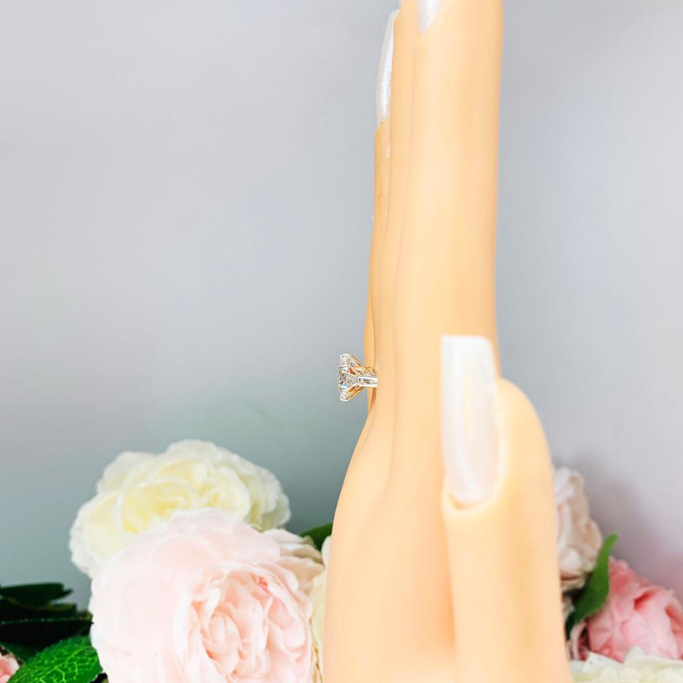 Mannequin hand with long white shimmery nails, displayed against a soft white-gray background with pink and white floral accents. The hand is wearing a sterling silver engagement ring with an oval lab created white sapphire set in a 6-prong tulip design setting.