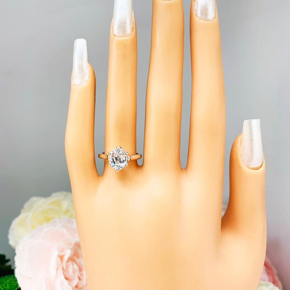 Mannequin hand with long white shimmery nails, displayed against a soft white-gray background with pink and white floral accents. The hand is wearing a sterling silver engagement ring with an oval lab created white sapphire set in a 6-prong tulip design setting.