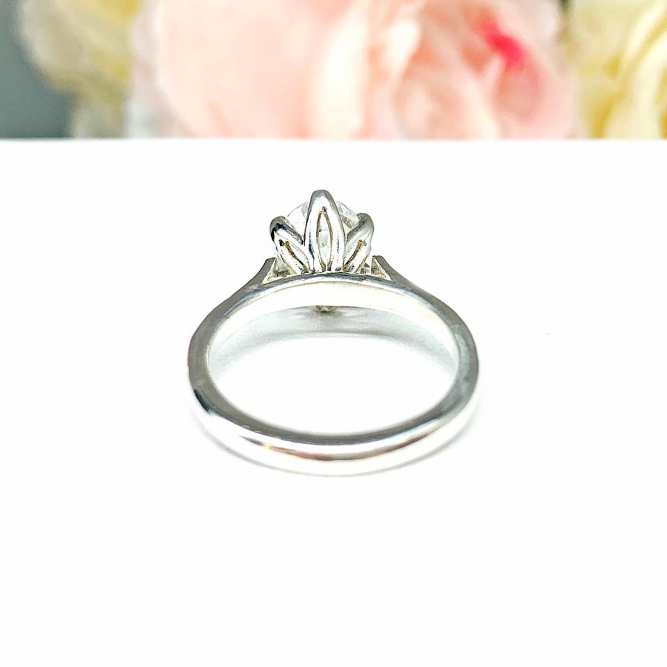 Sterling silver engagement ring with an oval lab created white sapphire set in a 6-prong tulip design setting set against a white surface with soft-focus pink and white flowers in the background.