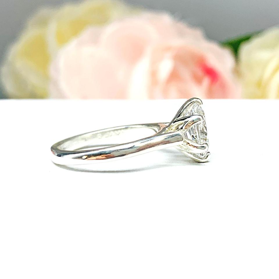 Sterling silver engagement ring with an oval lab created white sapphire set in a 6-prong tulip design setting set against a white surface with soft-focus pink and white flowers in the background.