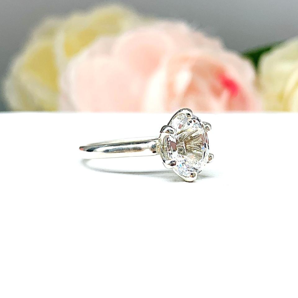 Sterling silver engagement ring with an oval lab created white sapphire set in a 6-prong tulip design setting set against a white surface with soft-focus pink and white flowers in the background.