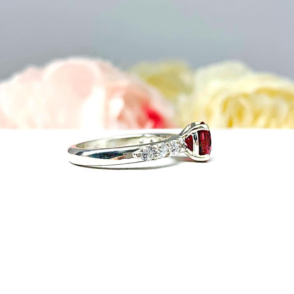 Sterling silver engagement ring with round lab-created deep red ruby and round CZ pave accents, shown on white surface.