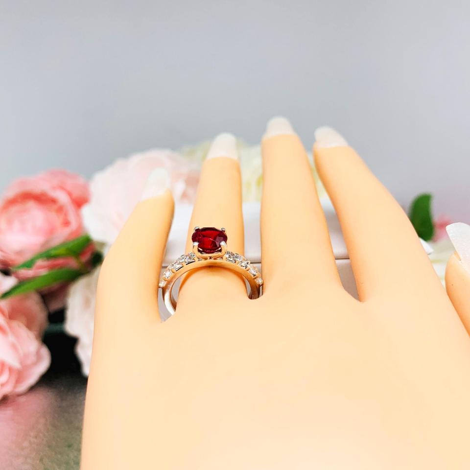 Mannequin hand with long white shimmery nails, displayed against a soft white-gray background with pink and white floral accents. The hand is wearing a sterling silver engagement ring with round lab-created deep red ruby and round CZ pave accents, shown on white surface.
