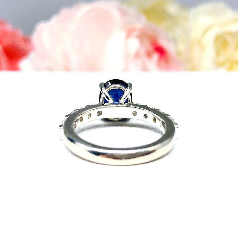 Close-up view of a stunning sterling silver oval-cut blue sapphire engagement ring. The engagement ring features a vibrant dark blue sapphire with pave cubic zirconia side stones.The ring is displayed against a blurred background of soft pink and white roses.