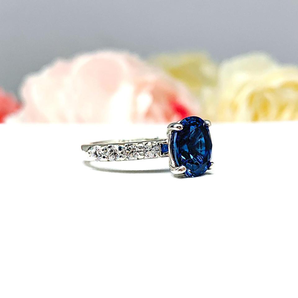 Close-up view of a stunning sterling silver oval-cut blue sapphire engagement ring. The engagement ring features a vibrant dark blue sapphire with pave cubic zirconia side stones.The ring is displayed against a blurred background of soft pink and white roses.