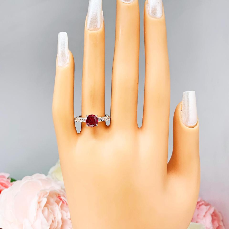 Mannequin hand with long white shimmery nails, displayed against a soft white-gray background with pink and white floral accents. The hand is wearing a sterling silver engagement ring with round lab-created deep red ruby and round CZ pave accents, shown on white surface.