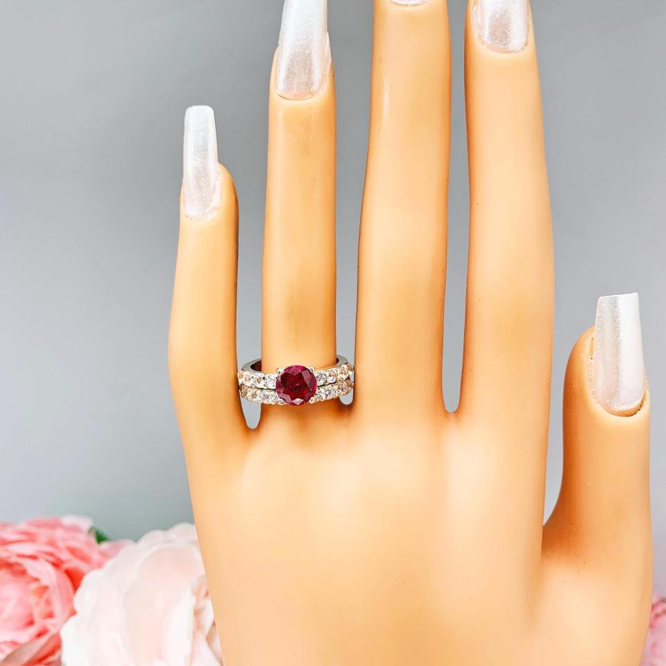 Mannequin hand with long white shimmery nails, displayed against a soft white-gray background with pink and white floral accents. The hand is wearing a sterling silver engagement ring with round lab-created deep red ruby and round CZ pave accents, shown with optional matching band.