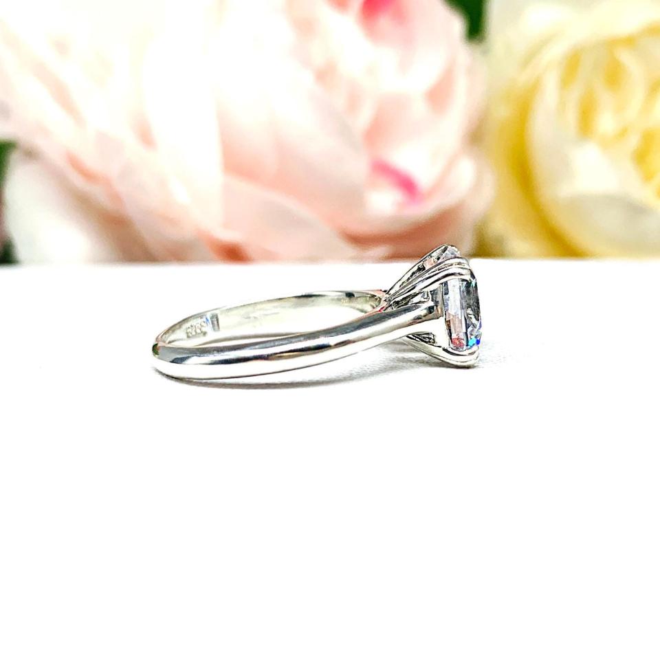 A sterling silver split-prong cathedral engagement ring set with a colorless old mine cut cubic zirconia. The ring is set against a white background with a blurred image of pink and white flowers in the background.