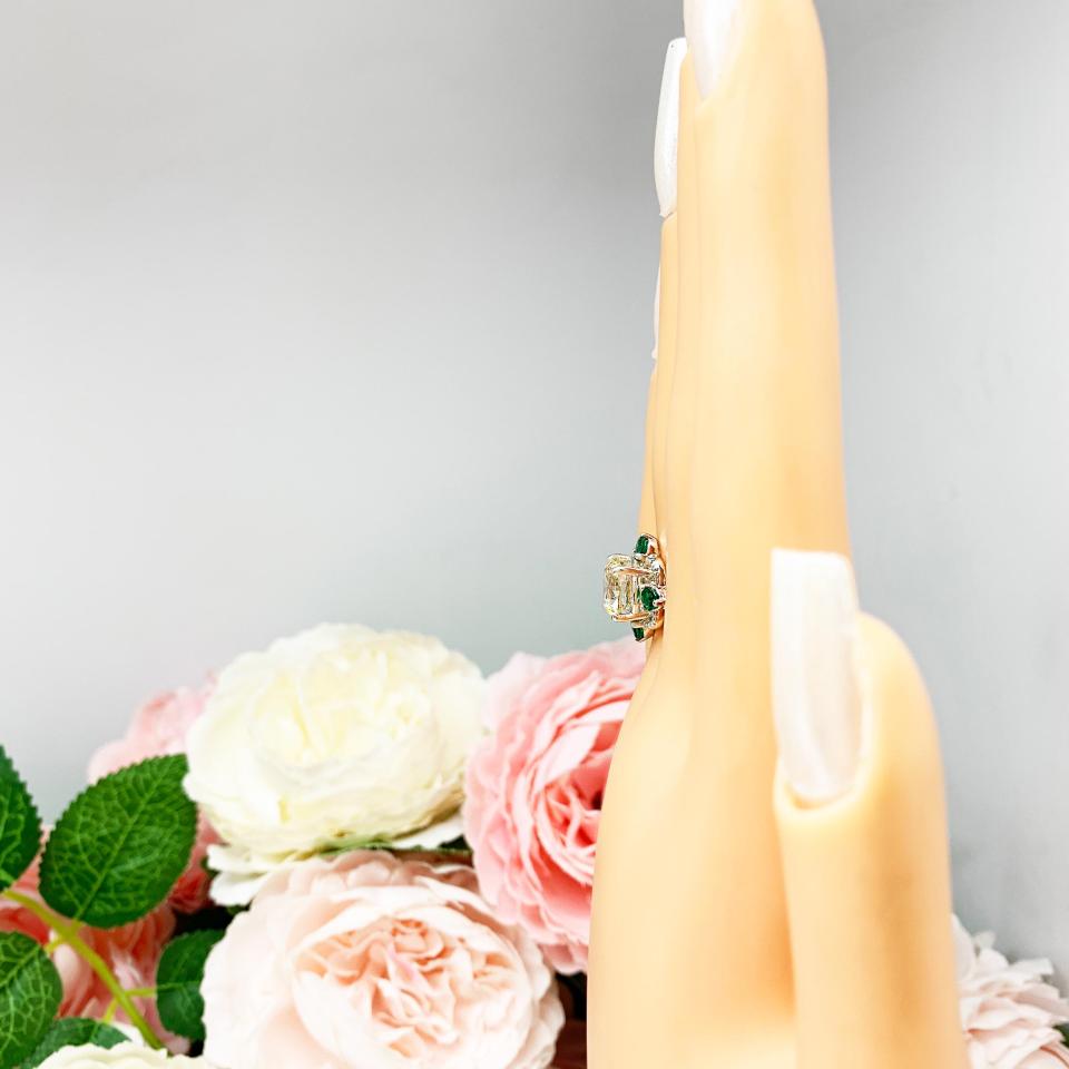 A display hand with long white fingernails standing up and with the top of the hand facing toward the left, displayed against a soft-focus background of pink and white flowers.  The display hand is wearing an Art Deco-style sterling silver ring with a round brilliant near-colorless cubic zirconia center stone, flanked by vivid green lab-created emerald accents.