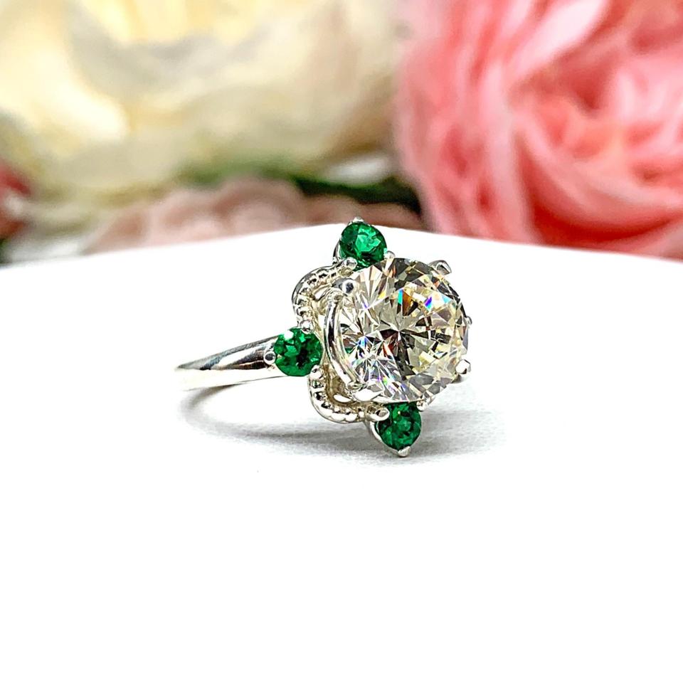 Art Deco-style sterling silver ring with a round brilliant near-colorless cubic zirconia center stone, flanked by vivid green lab-created emerald accents. The ring is facing slightly to the right and is photographed sitting on a white surface with background of pink and white flowers.