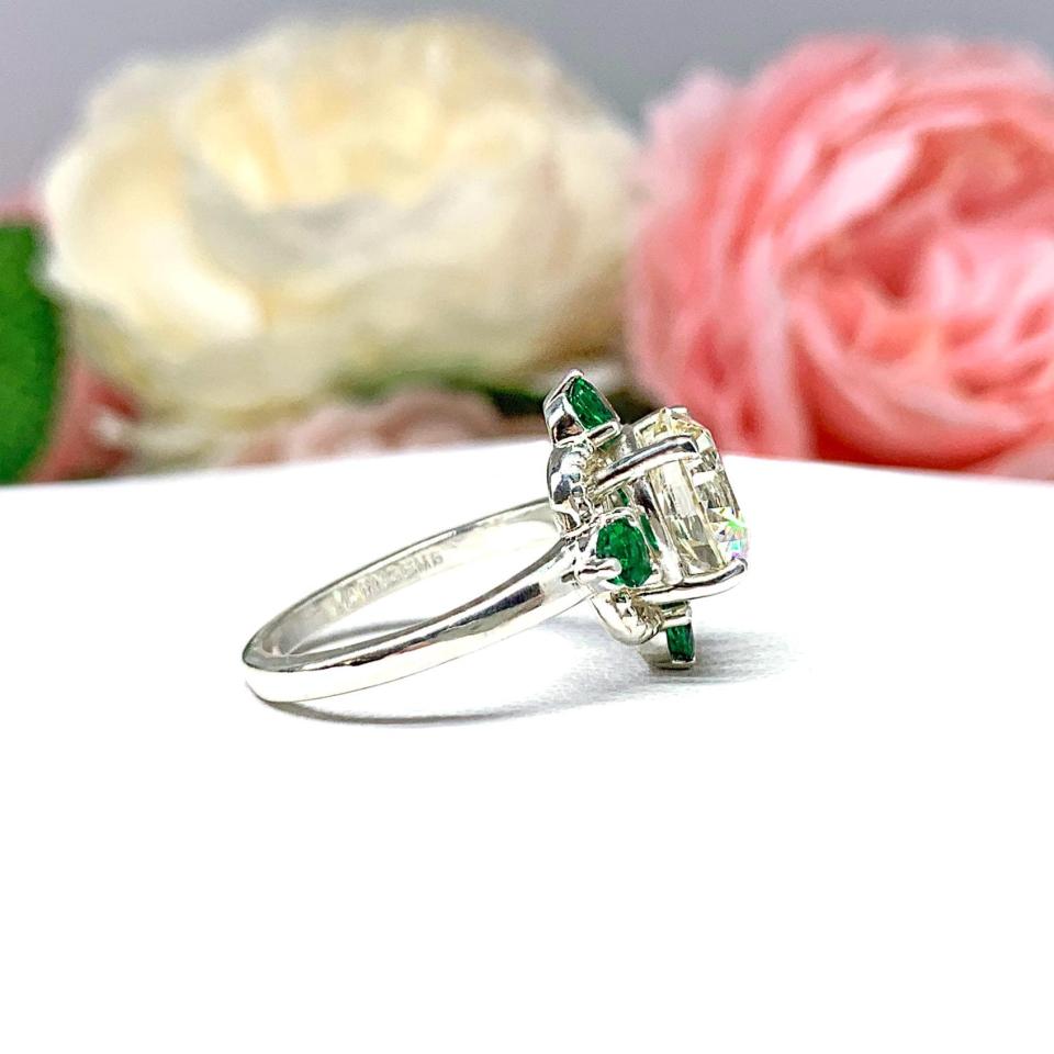 Art Deco-style sterling silver ring with a round brilliant near-colorless cubic zirconia center stone, flanked by vivid green lab-created emerald accents. The ring is facing toward the right and is photographed sitting on a white surface with background of pink and white flowers.