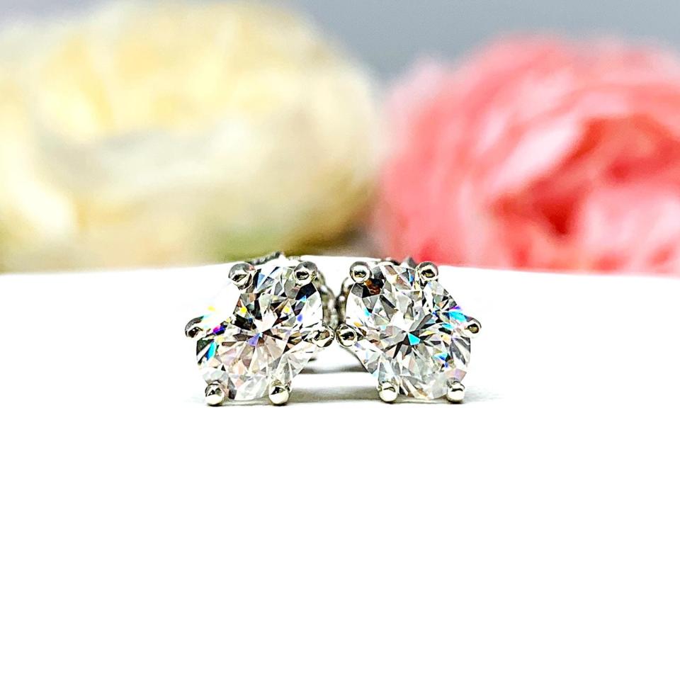 A pair of 10k white gold stud earrings set with oval brilliant cut moissanites.  The 6 prong earrings are on a white surface with both earrings facing slightly to the right to showcase the brilliant cutting of the moissanite, center post and butterfly friction clasps.
