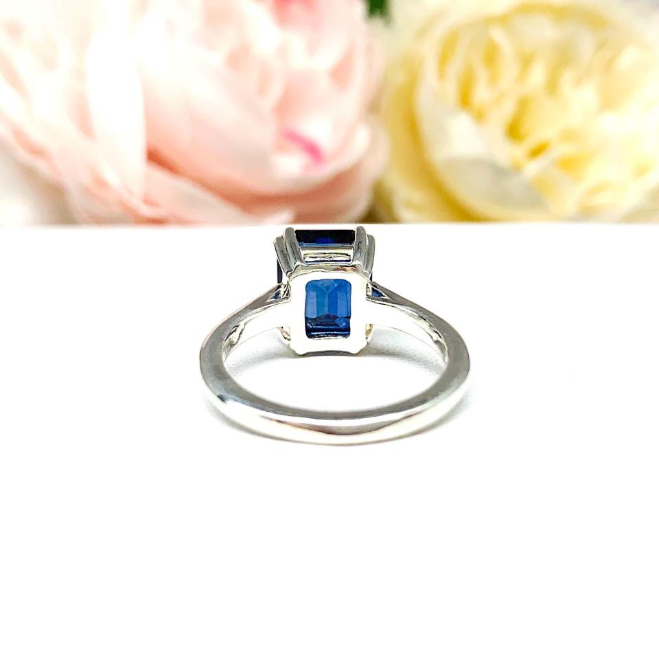 Sterling silver engagement ring featuring a deep blue emerald-cut lab-created blue sapphire in a sleek double-prong setting. The ring is resting on a white surface with soft-focus pink and white flowers.
