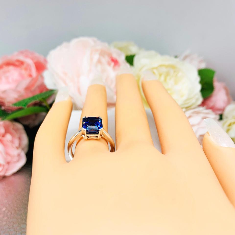 Mannequin hand with long white shimmery nails, displayed against a soft white-gray background with pink and white floral accents. The hand is wearing a sterling silver solitaire ring featuring a deep blue emerald-cut lab-created blue sapphire in a sleek double-prong setting. The text '10X8MM' appears in the image.