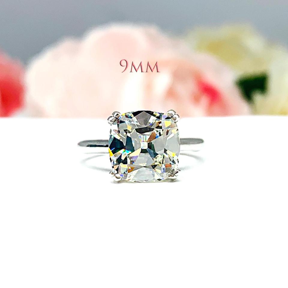 A sterling silver split-prong cathedral engagement ring set with a near colorless old mine cut cubic zirconia. The ring is set against a white background with a blurred image of pink and white flowers in the background. The text '9MM' appears in the image.