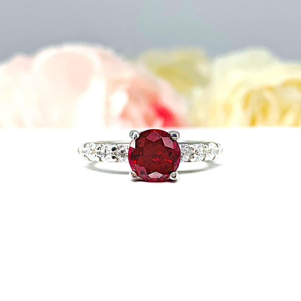 Sterling silver engagement ring with round lab-created deep red ruby and round CZ pave accents, shown on white surface.