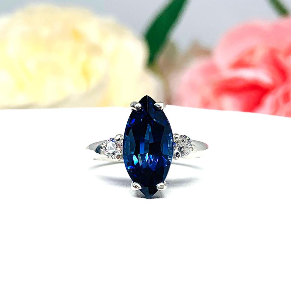 Sterling silver ring showcasing a striking marquise-cut lab-created blue sapphire, it is flanked by two brilliant round cubic zirconia stones. The ring is set against a soft floral backdrop that enhances the ring’s romantic allure.