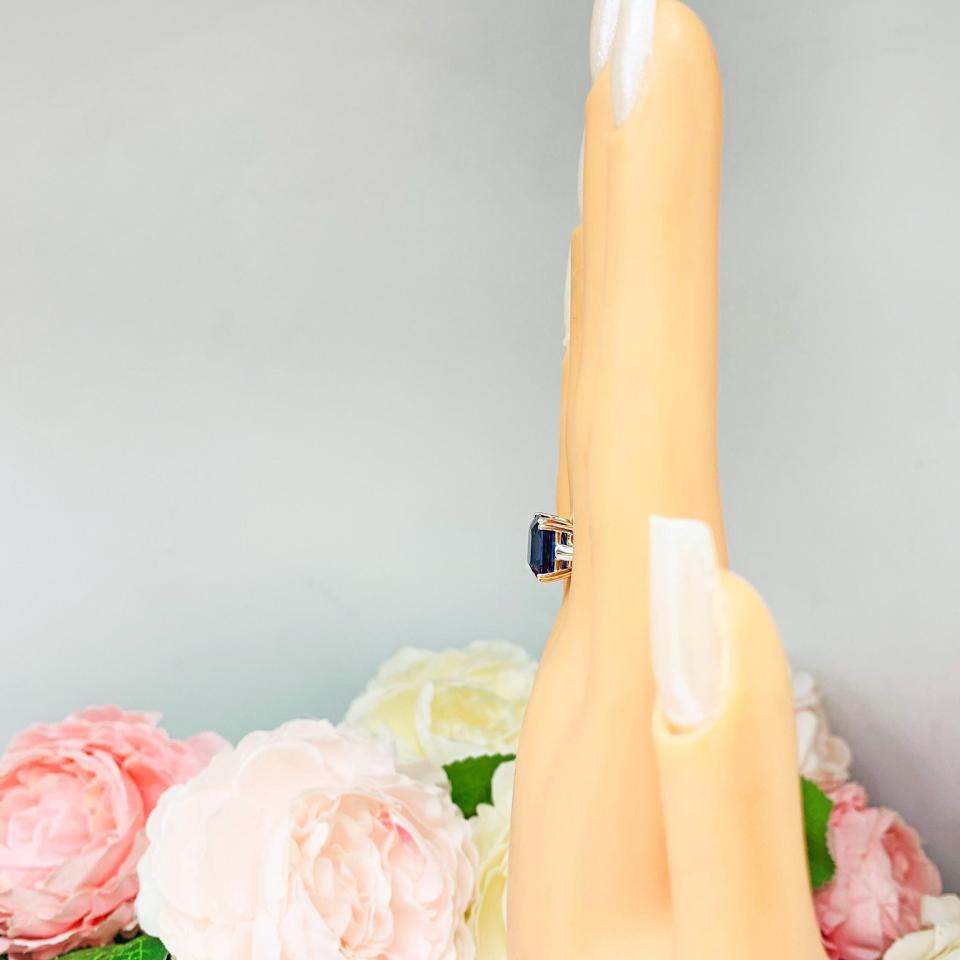 Mannequin hand with long white shimmery nails, displayed against a soft white-gray background with pink and white floral accents. The hand is wearing a sterling silver solitaire ring featuring a deep blue emerald-cut lab-created blue sapphire in a sleek double-prong setting. The text '10X8MM' appears in the image.