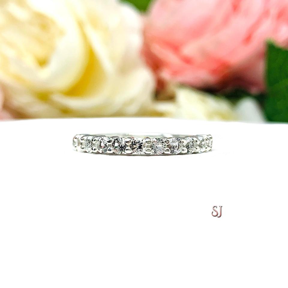 Sterling silver band with 12 round 2.5mm white topaz gemstones set in a shared prong setting. The band is on a white surface with pink and white flowers in the background.