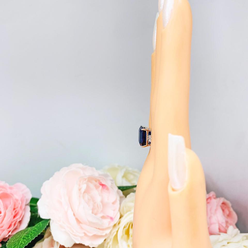 Mannequin hand with long white shimmery nails, displayed against a soft white-gray background with pink and white floral accents. The hand is wearing a sterling silver three stone ring featuring a deep blue emerald cut lab created blue sapphire flanked by a round brilliant cz on each side.