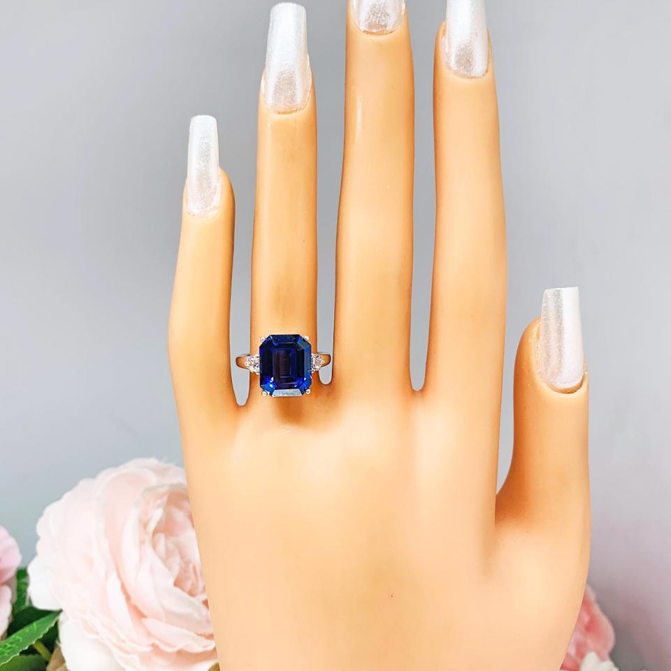 Mannequin hand with long white shimmery nails, displayed against a soft white-gray background with pink and white floral accents. The hand is wearing a sterling silver three stone ring featuring a deep blue emerald cut lab created blue sapphire flanked by a round brilliant cz on each side.