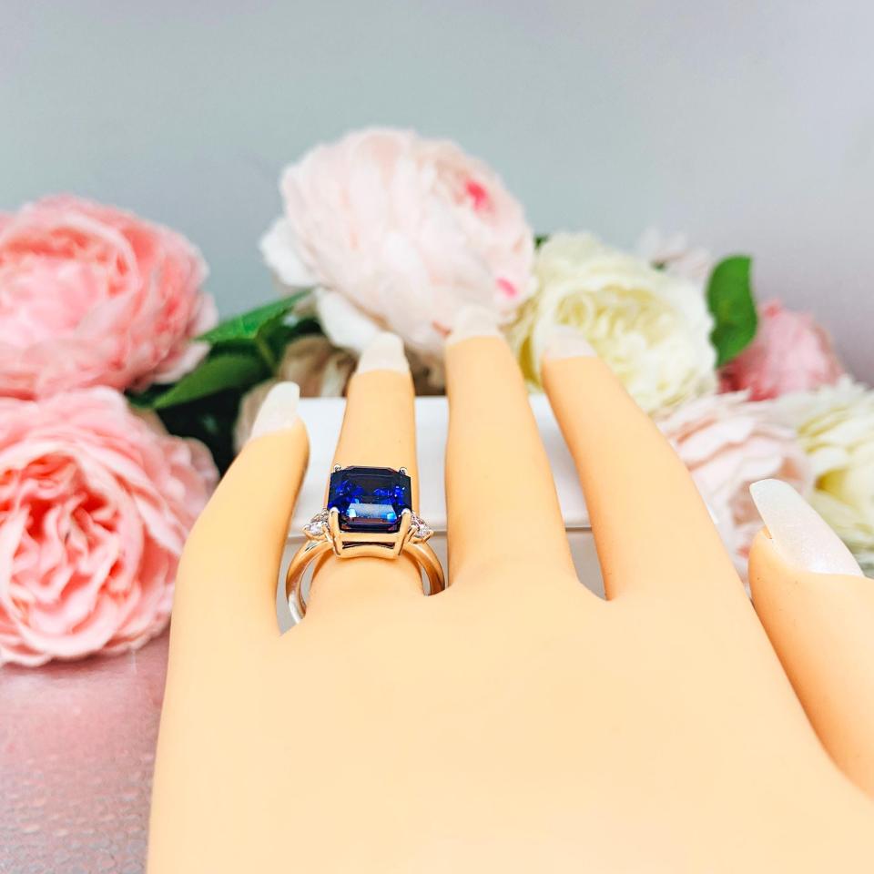 Mannequin hand with long white shimmery nails, displayed against a soft white-gray background with pink and white floral accents. The hand is wearing a sterling silver three stone ring featuring a deep blue emerald cut lab created blue sapphire flanked by a round brilliant cz on each side.