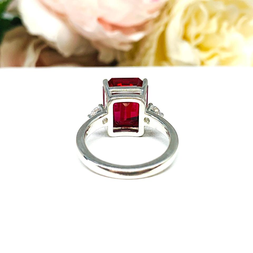 A sterling silver three stone ring featuring a deep red emerald cut lab created ruby flanked by a round brilliant cz on each side. The ring is resting on a white surface with soft-focus pink and white flowers in the background.