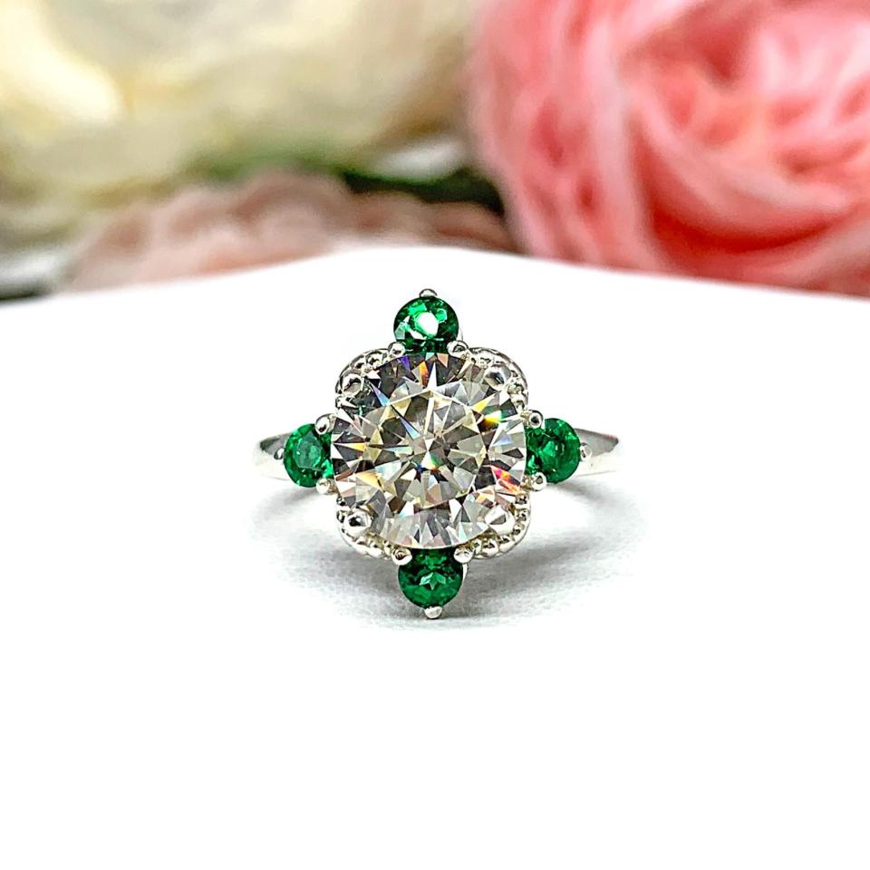 Art Deco-style sterling silver ring with a round brilliant near-colorless cubic zirconia center stone, flanked by vivid green lab-created emerald accents. The ring is photographed sitting on a white surface with background of pink and white flowers.
