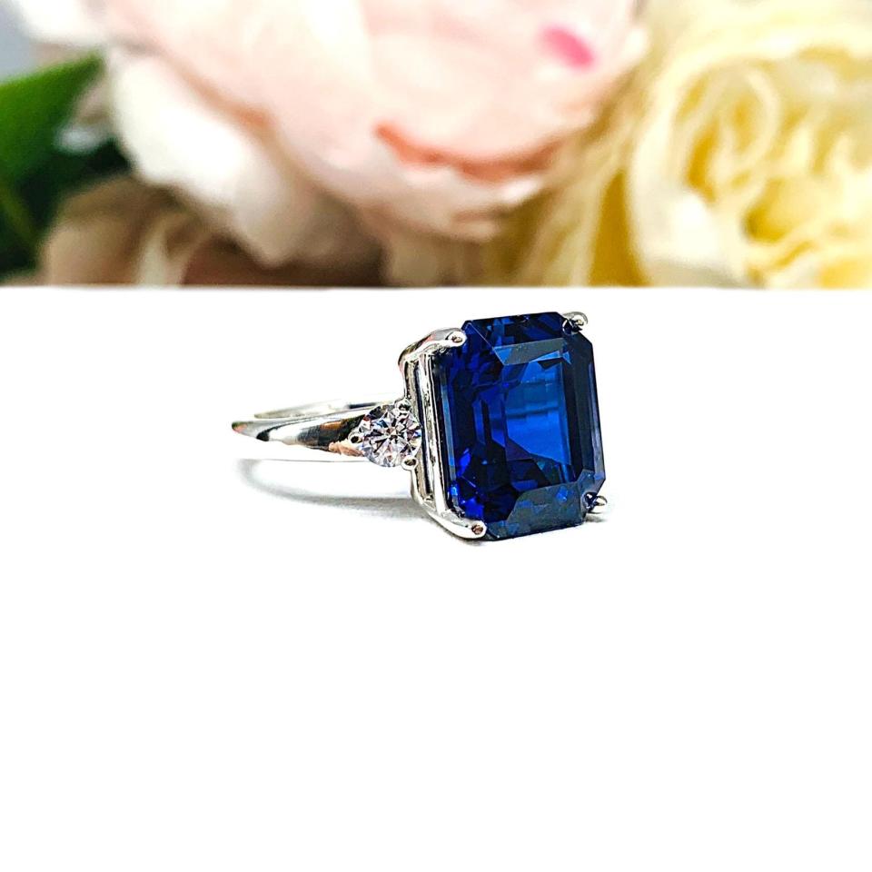 A sterling silver three stone ring featuring a deep blue emerald cut lab created blue sapphire flanked by a round brilliant cz on each side. The ring is resting on a white surface with soft-focus pink and white flowers in the background.