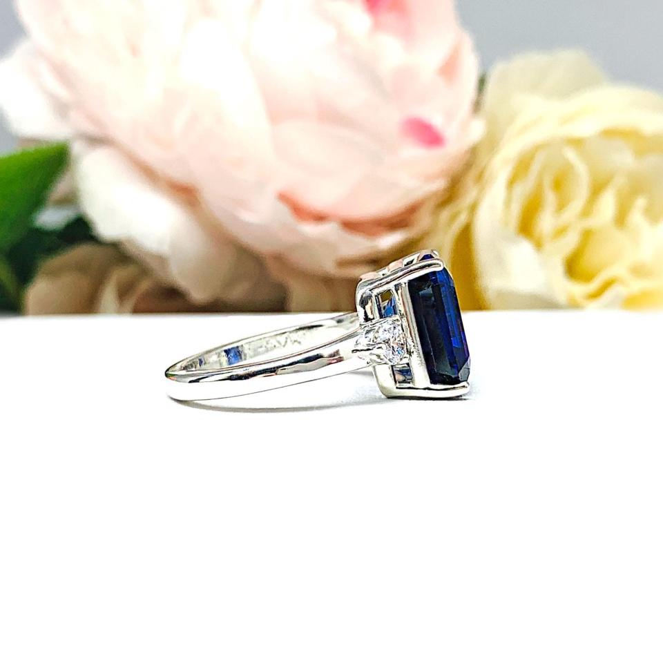 A sterling silver three stone ring featuring a deep blue emerald cut lab created blue sapphire flanked by a round brilliant cz on each side. The ring is resting on a white surface with soft-focus pink and white flowers in the background.