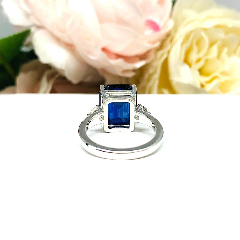 A sterling silver three stone ring featuring a deep blue emerald cut lab created blue sapphire flanked by a round brilliant cz on each side. The ring is resting on a white surface with soft-focus pink and white flowers in the background.