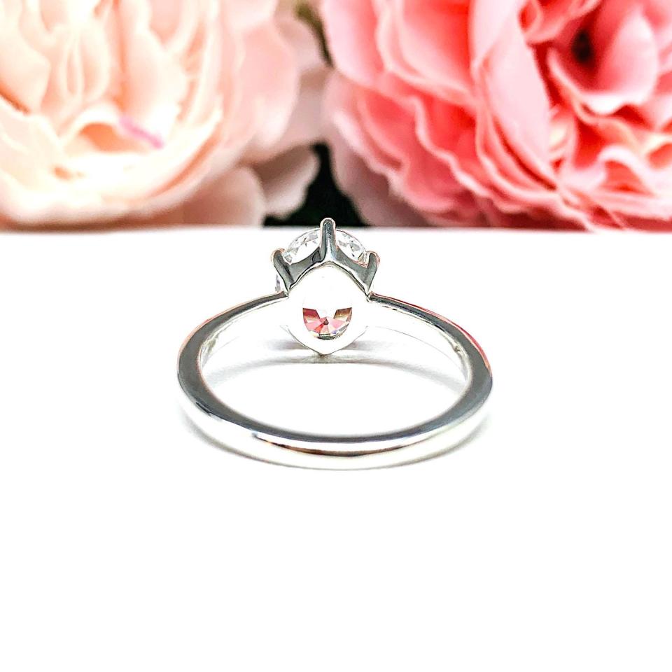 Sterling silver ring with an oval lab created white sapphire set in a 6-prong setting set against a white surface with soft-focus pink flowers in the background.