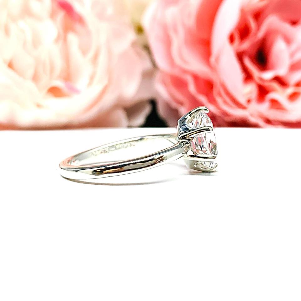 Sterling silver ring with an oval lab created white sapphire set in a 6-prong setting set against a white surface with soft-focus pink flowers in the background.