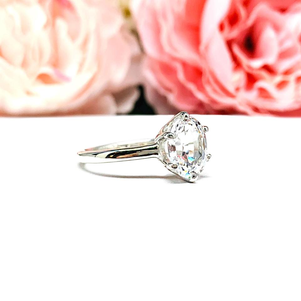 Sterling silver ring with an oval lab created white sapphire set in a 6-prong setting set against a white surface with soft-focus pink flowers in the background.