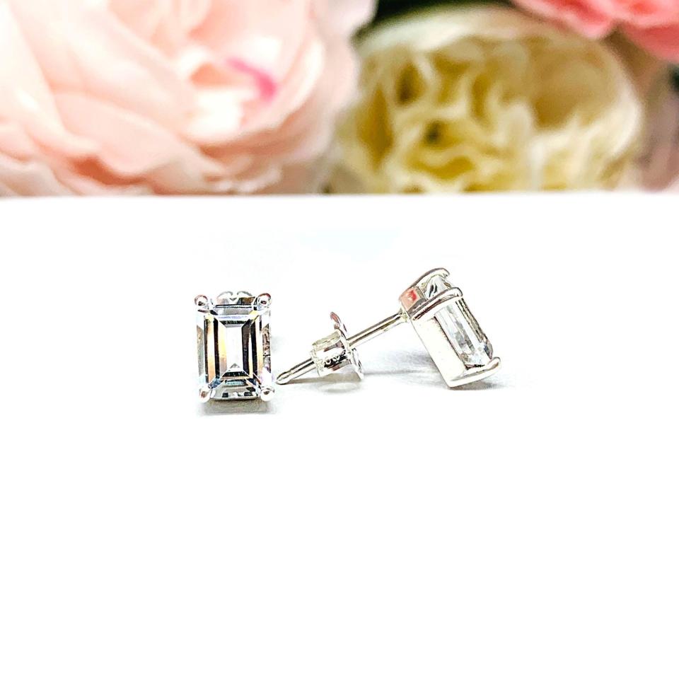 A pair of silver earrings set with emerald cut natural white topaz gemstones, displayed with one earring facing the front to showcase clear clarity of the gemstones and one facing to the side to display that the post sits at the top of the earring and the butterfly friction clasp. The earrings are shown on a white background.
