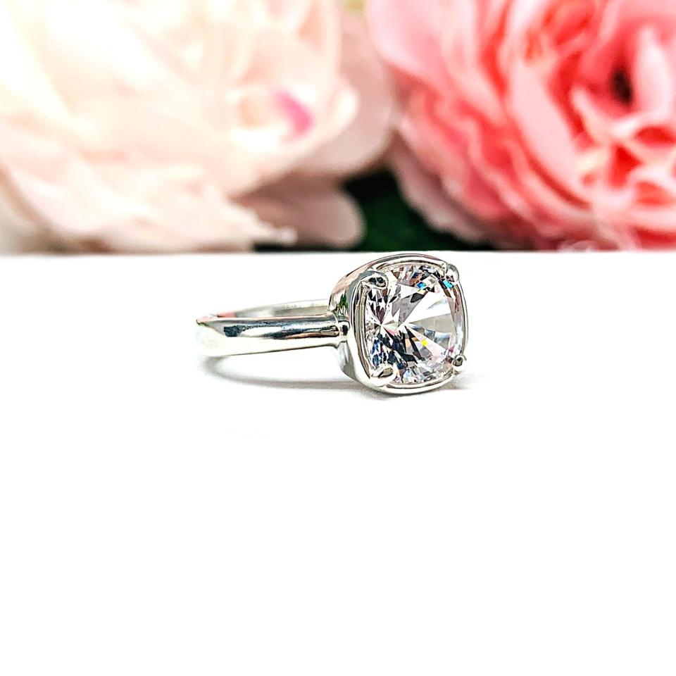 A sterling silver ring with a large, cushion-cut lab white sapphire. The stone is set in a bezel prong setting, and the band is rounded and polished. The background features soft pink and white flowers, creating a romantic aesthetic.