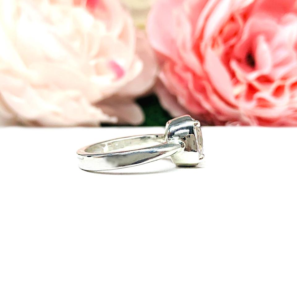 A sterling silver ring with a large, cushion-cut lab white sapphire. The stone is set in a bezel prong setting, and the band is rounded and polished. The background features soft pink and white flowers, creating a romantic aesthetic.