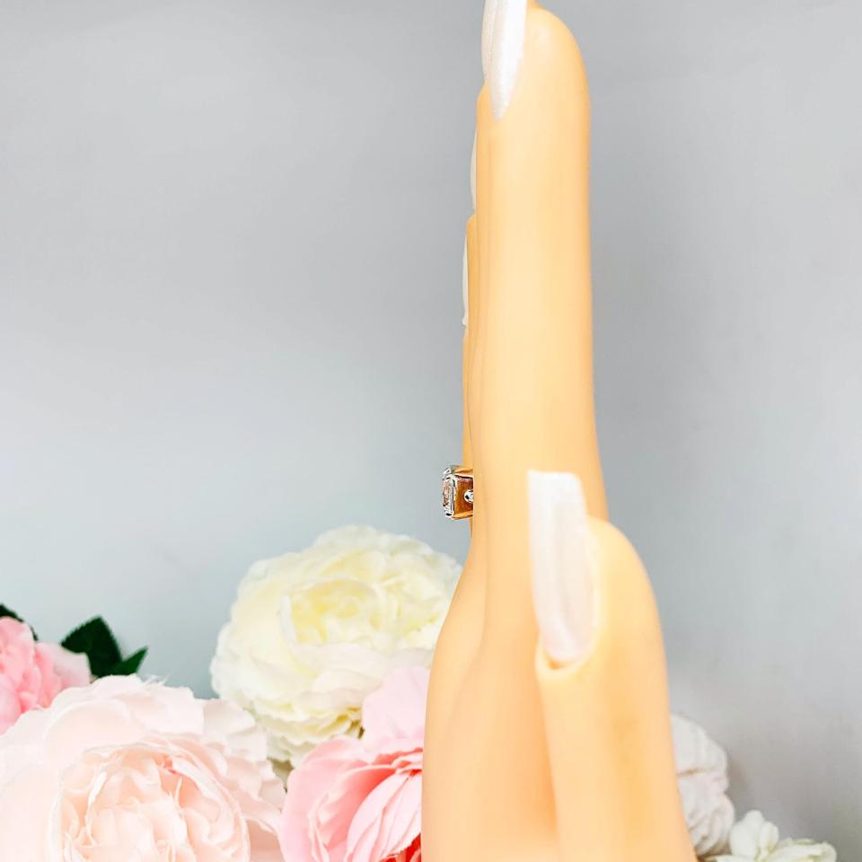 Mannequin hand with long white shimmery nails, displayed against a soft white-gray background with pink and white floral accents. The hand is wearing a sterling silver ring with a large, cushion-cut lab white sapphire. The stone is set in a bezel prong setting, and the band is rounded and polished.