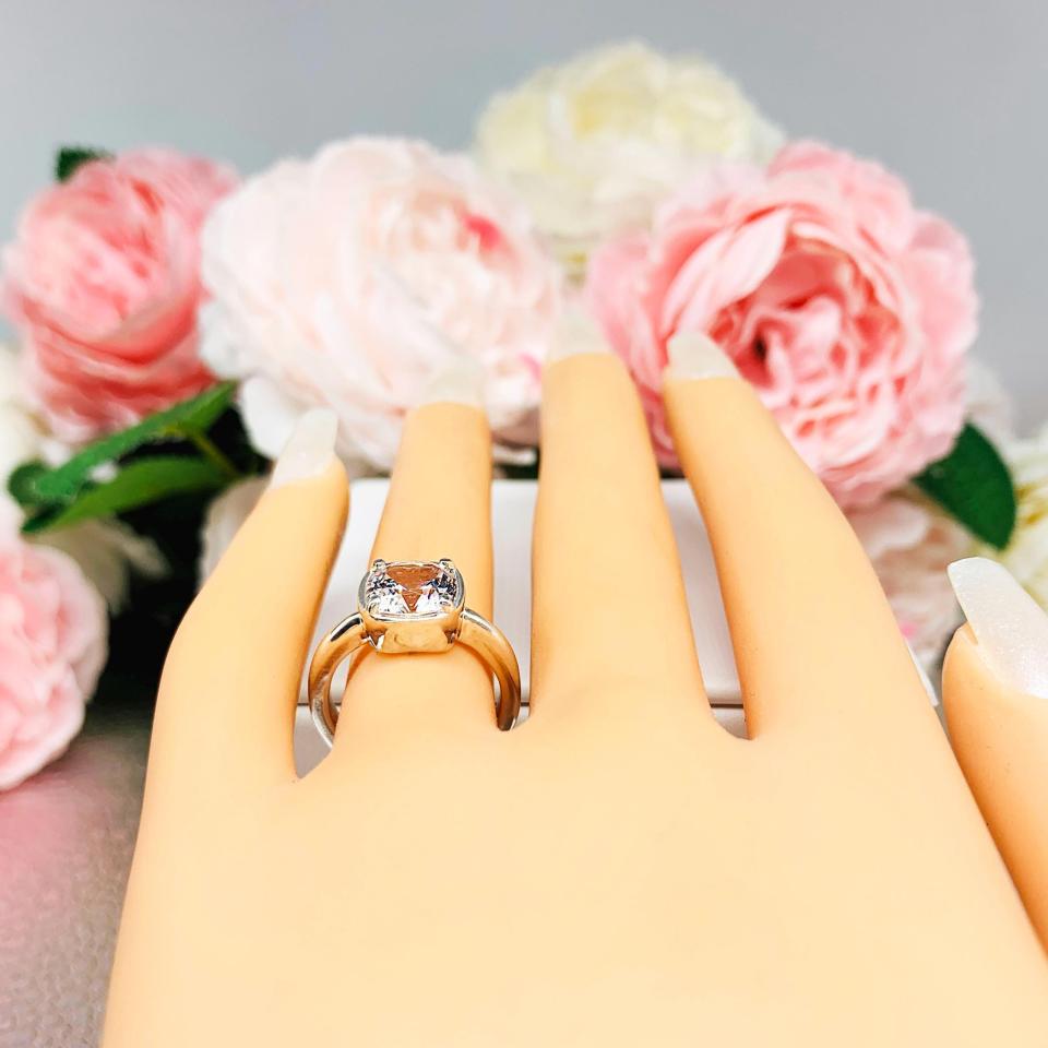 Mannequin hand with long white shimmery nails, displayed against a soft white-gray background with pink and white floral accents. The hand is wearing a sterling silver ring with a large, cushion-cut lab white sapphire. The stone is set in a bezel prong setting, and the band is rounded and polished.