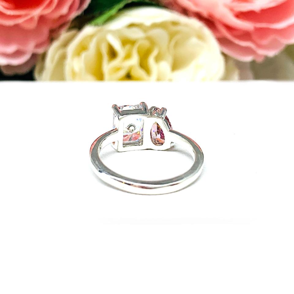Close‑up of a sterling silver toi et moi ring featuring a an 8x5mm morganite pink color pear‑cut CZ and an 8x6mm colorless radiant-cut cz. Displayed on a white surface with a floral background.
