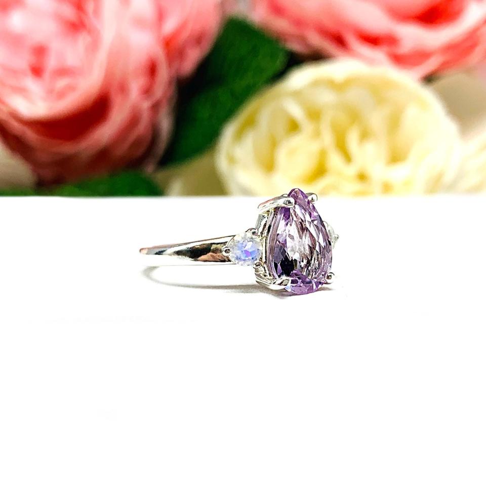 Sterling silver ring with a pear‑cut Rose de France amethyst and round rainbow moonstones. Displayed on a white surface with soft pastel floral background.