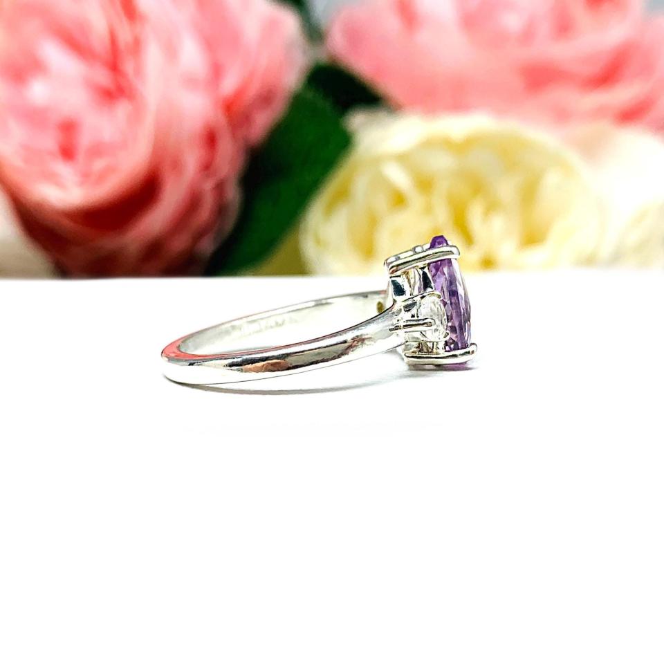 Sterling silver ring with a pear‑cut Rose de France amethyst and round rainbow moonstones. Displayed on a white surface with soft pastel floral background.