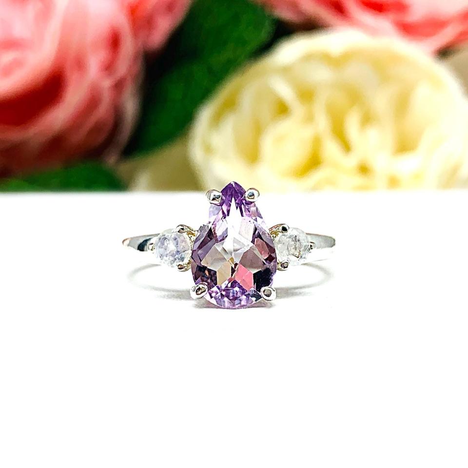 Sterling silver ring with a pear‑cut Rose de France amethyst and round rainbow moonstones. Displayed on a white surface with soft pastel floral background.