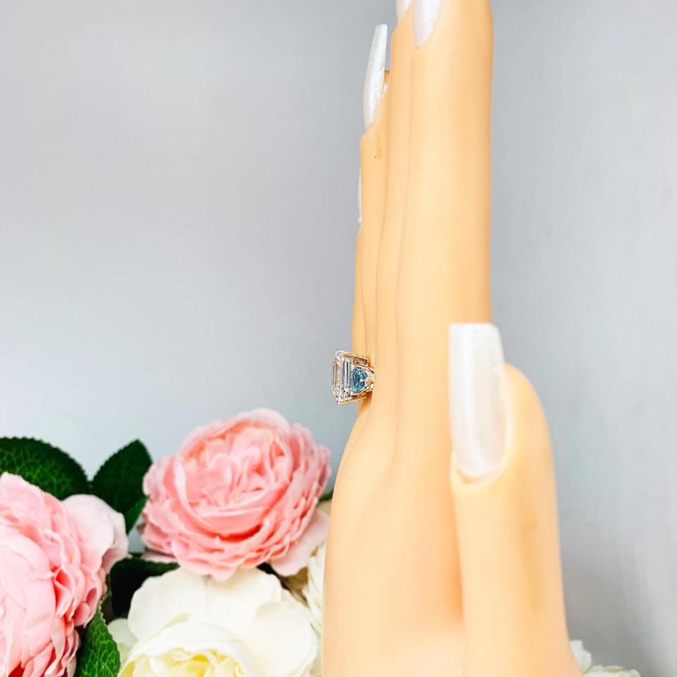 Mannequin hand with long white shimmery nails, displayed against a soft white-gray background with pink and white floral accents. The hand is wearing a sterling silver 3-stone ring featuring an 11x9mm lab created white sapphire flanked by two 5mm swiss blue topaz gemstones.